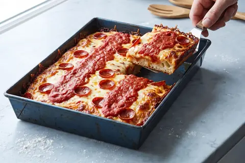 A rectangular pizza in a dark steel pan: caramelised cheese along the edges, stripes of tomato sauce, pepperoni, and one square lifted out on a spatula.