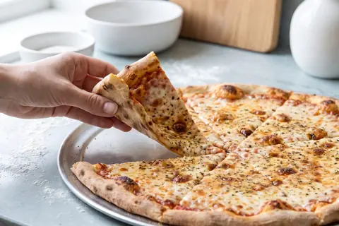 A large, thin pizza with melted cheese and oregano on a metal tray; a hand lifts a wide slice.